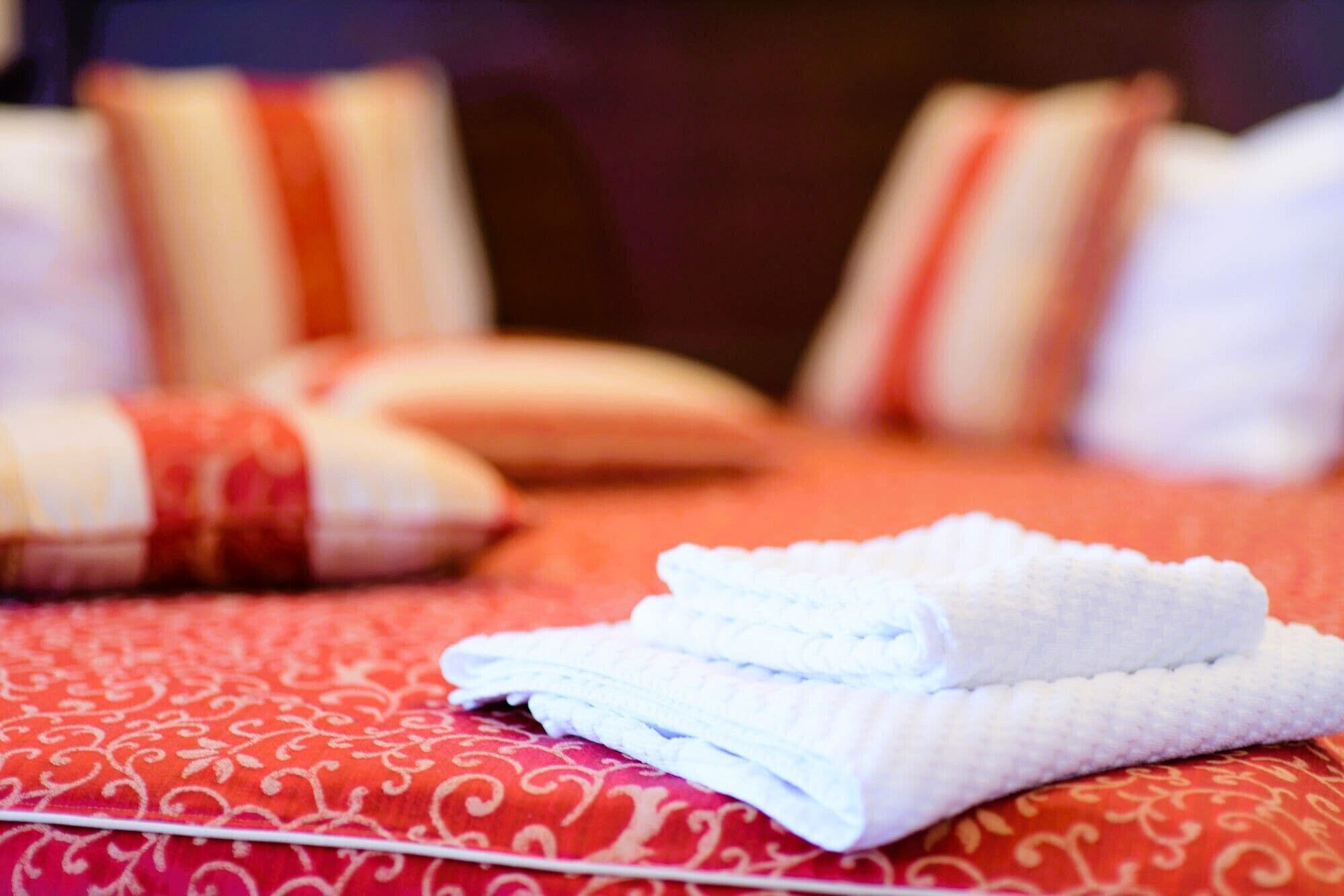 A neatly folded white towel on a patterned bedspread with pillows in the background.