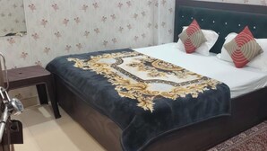 A neatly made bed with a patterned bedspread and decorative pillows, a bedside table, and a chair in a room with floral wallpaper. - Shree Radharaman Palace (Ayodhya)