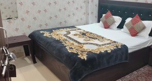 A neatly made bed with a patterned bedspread and decorative pillows, a bedside table, and a chair in a room with floral wallpaper. - Shree Radharaman Palace (Ayodhya)