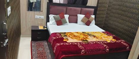 A neatly made bed with a red and yellow floral bedspread, a wooden nightstand with a lamp, and a red patterned rug.