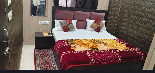 A neatly made bed with a red and yellow floral bedspread, a wooden nightstand with a lamp, and a red patterned rug. - Shree Radharaman Palace (Ayodhya)