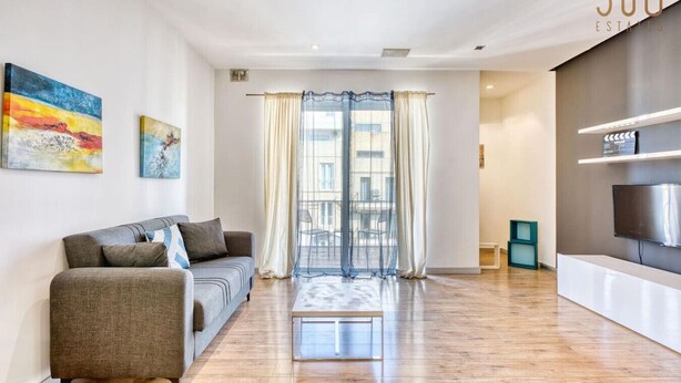 Ein modernes Wohnzimmer mit einer Couch, einem Couchtisch und einem Fernseher. Ein großes Fenster mit Vorhängen und Kunstwerken an den Wänden.