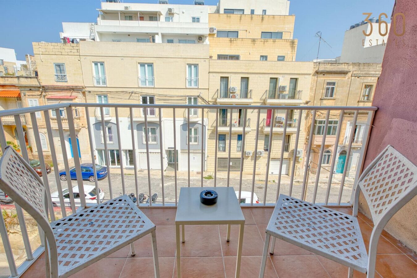 Un balcon avec deux chaises en métal et une petite table, donnant sur un quartier résidentiel composé de plusieurs immeubles.