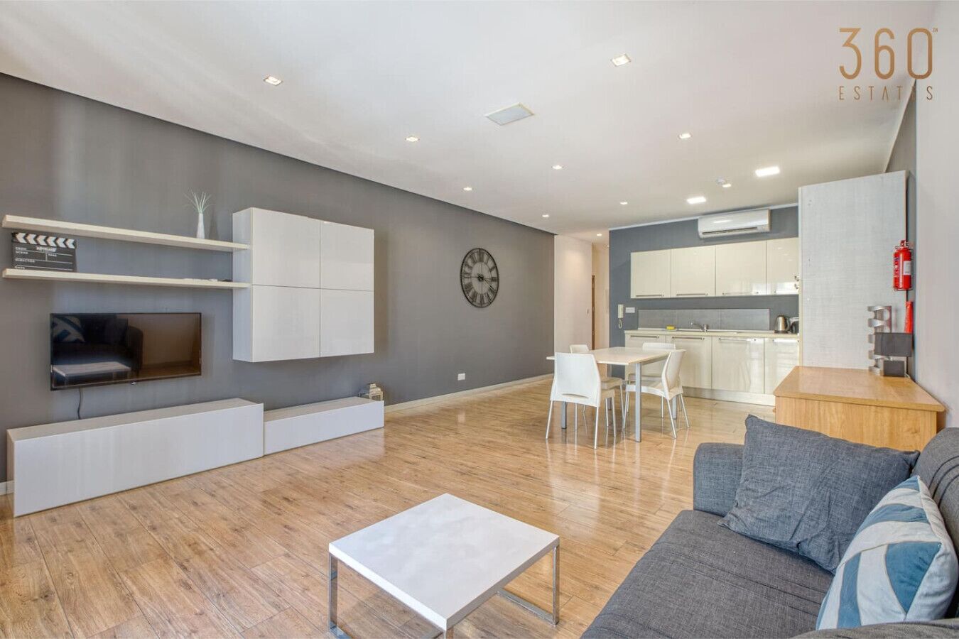 Un salon moderne doté d’un téléviseur à écran plat, d’étagères intégrées, d’un coin repas avec une table et des chaises, et d’une cuisine en arrière-plan.