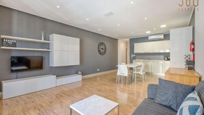 Ein modernes Wohnzimmer mit Flachbildfernseher, Einbauschränken, einem Essbereich mit Tisch und Stühlen sowie einer Küche im Hintergrund.