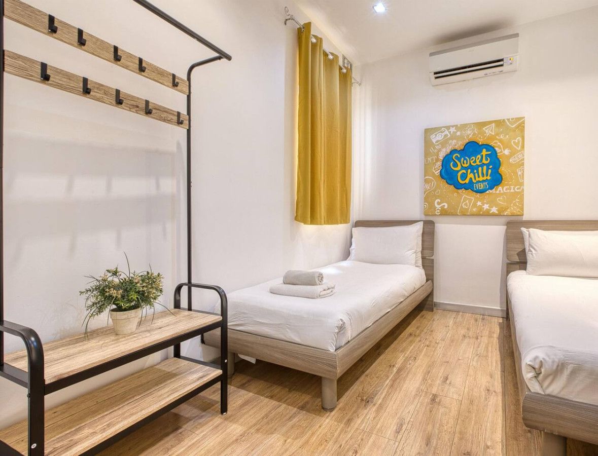 A hotel room with two beds, a small table with a plant, a wall-mounted air conditioner, and a 'Sweet Chill Events' sign.