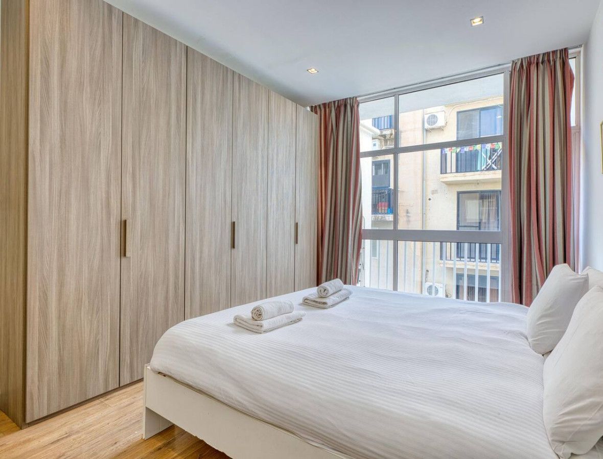 Une chambre avec un grand lit, une armoire, une fenêtre avec des rideaux et une vue sur les bâtiments environnants.