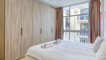 Une chambre avec un grand lit, une armoire, une fenêtre avec des rideaux et une vue sur les bâtiments environnants.