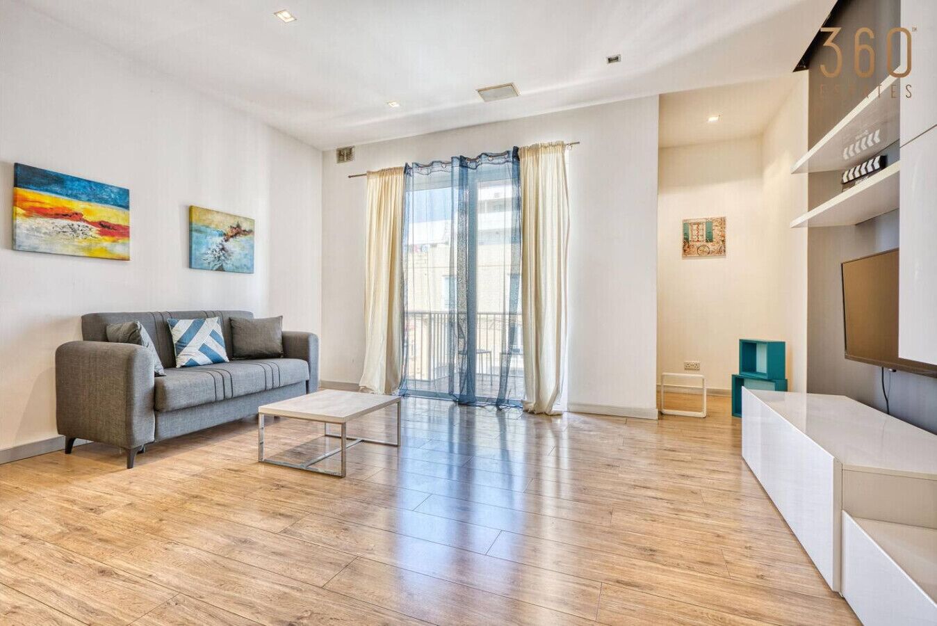 Un salon moderne avec un canapé gris, une table basse et un téléviseur posé sur un meuble blanc. Deux tableaux ornent le mur et un balcon est agrémenté de rideaux.