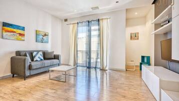 Un salon moderne avec un canapé gris, une table basse et un téléviseur posé sur un meuble blanc. Deux tableaux ornent le mur et un balcon est agrémenté de rideaux.