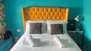 Un letto rifatto con lenzuola bianche e due cuscini grigi, una luminosa testata imbottita arancione e un comodino con una lampada e un cartello con scritto 'Guest Information'.