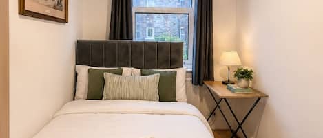 A hotel room with a bed, a bedside table with a lamp, a window with curtains, and a painting on the wall.
