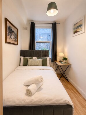 A hotel room with a bed, a bedside table with a lamp, a window with curtains, and a painting on the wall. - The Cozy Settle Stay - Relax and Unwind, 2 bedroom apartment near central London (London)