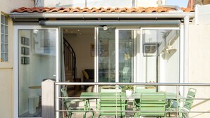 Un patio cubierto con sillas de metal verde y una mesa, una puerta corrediza de vidrio que da a un espacio interior, y un techo de tejas.