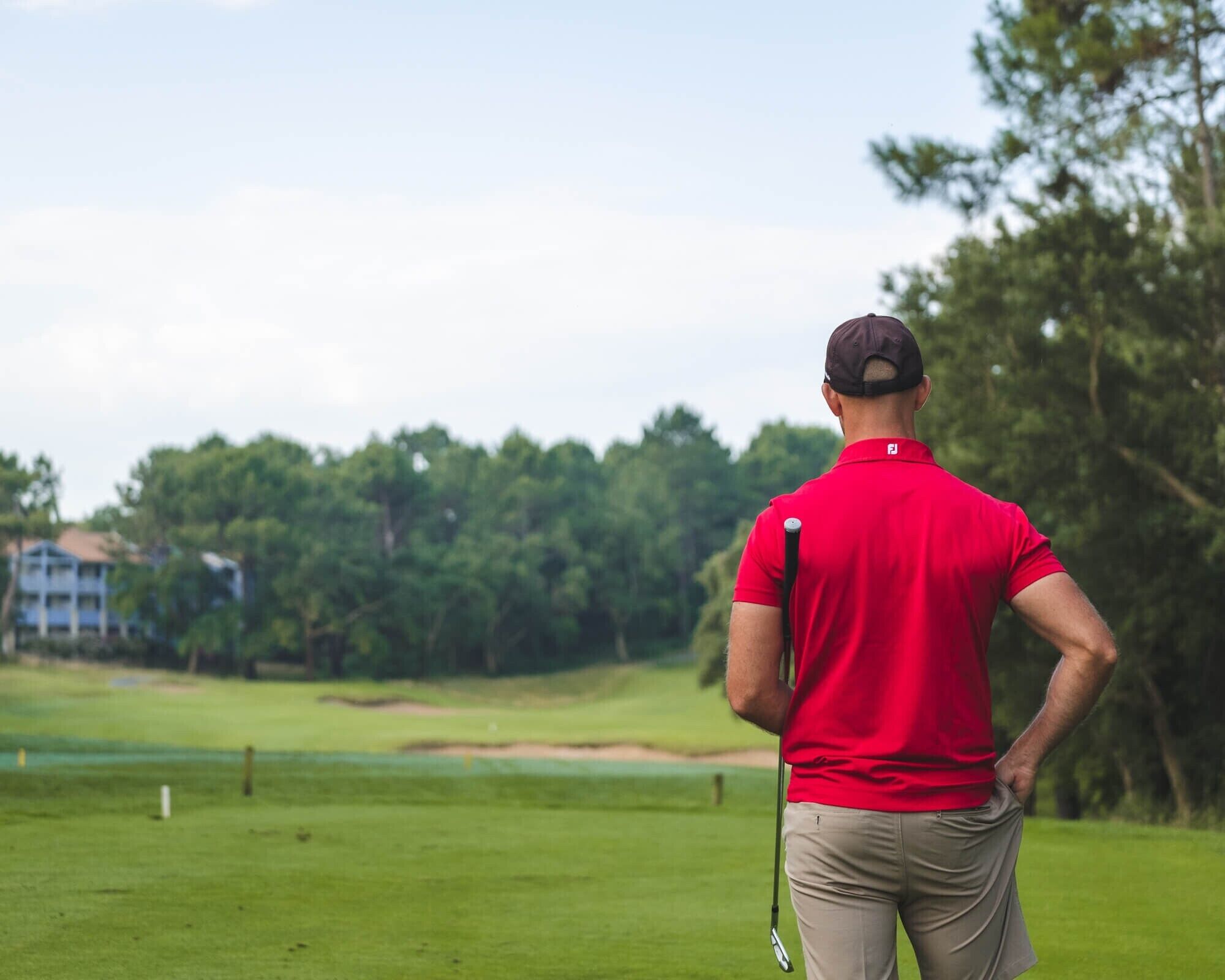 Una persona con camisa y gorra roja, de pie en un campo de golf con un palo de golf, mirando alejándose de la cámara.