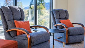 Two grey armchairs with wooden armrests and orange cushions, a small black table, and a view of trees and mountains through large windows.
