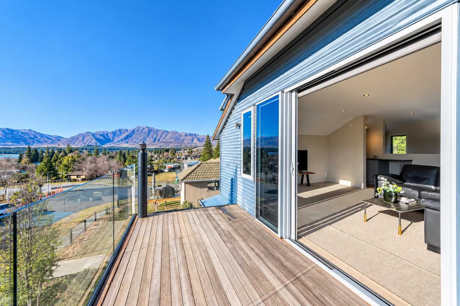 A modern balcony with a wooden deck, a sliding glass door leading to a living area with a sofa and a coffee table, and a scenic mountain view in the background.