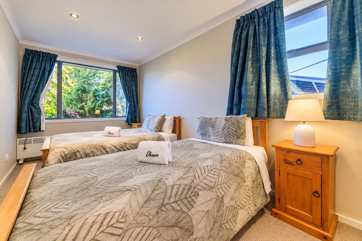 A hotel room with two beds, a wooden nightstand, a lamp, and a window with curtains.