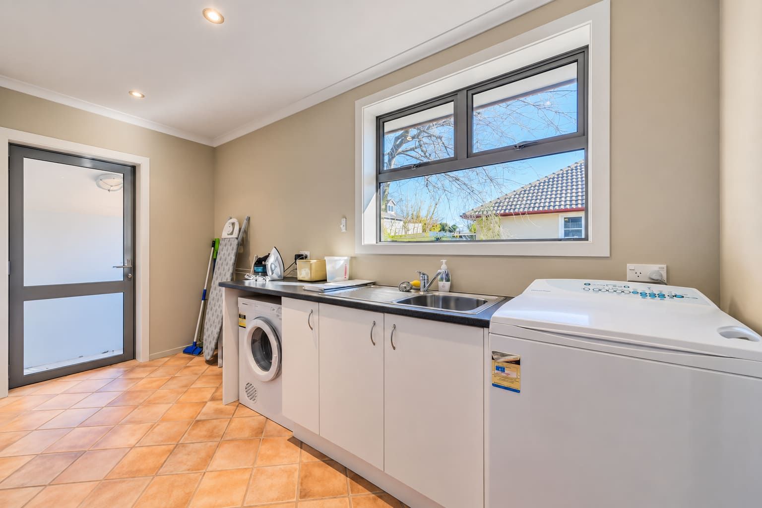 A laundry room with a washing machine, a dryer, a sink, and a window with a view of a house.