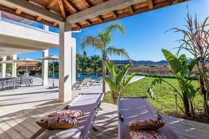 Un espacio al aire libre techado con sillones de descanso y una piscina, rodeado de vegetación y una vista despejada del paisaje.