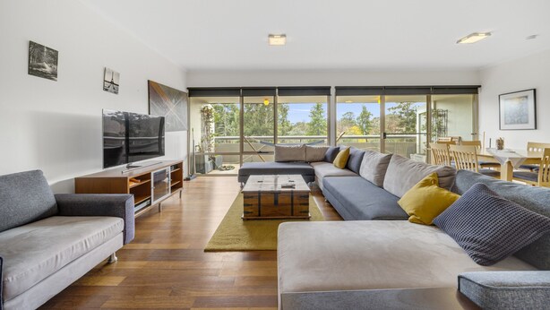 A modern living room with a large sofa, a coffee table, and a TV. There are large windows offering a view of the outdoors. - Bright 2-bed With Private Balcony (Canberra)