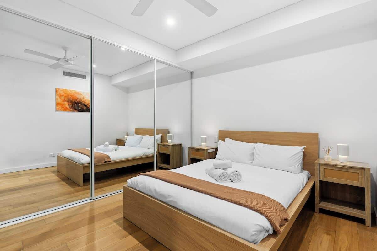 A modern bedroom with a large bed, two bedside tables, a ceiling fan, and a glass partition.