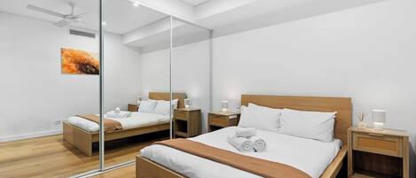 A modern bedroom with a large bed, two bedside tables, a ceiling fan, and a glass partition.