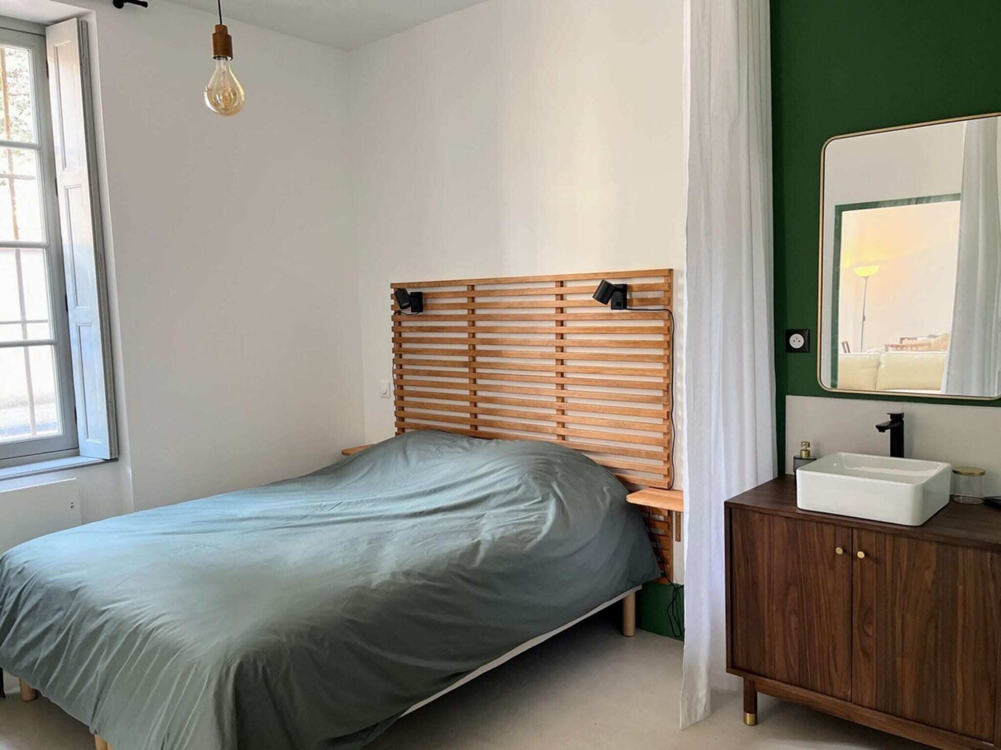 Une chambre à coucher avec un lit, une tête de lit en bois, une table de chevet avec un miroir et une fenêtre avec des rideaux.