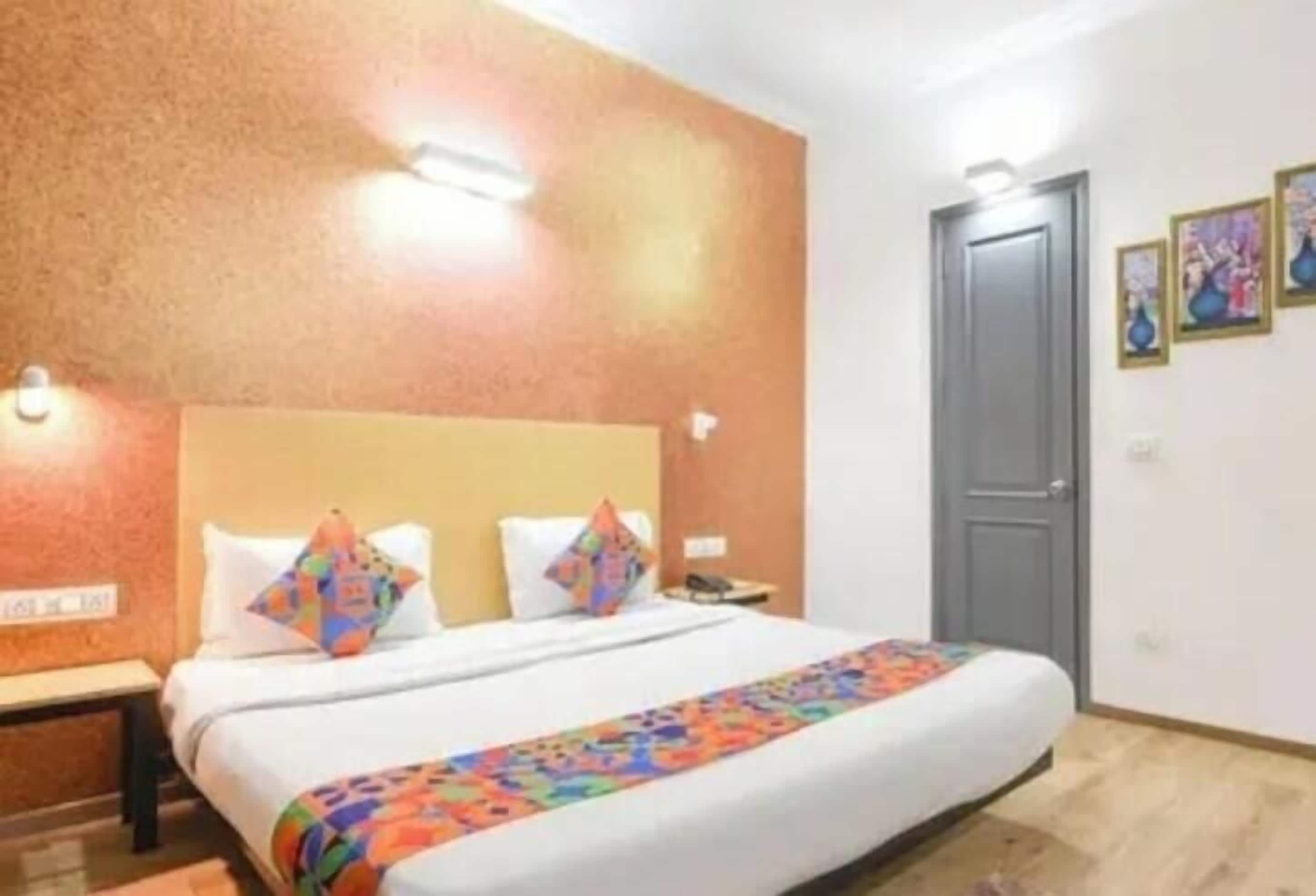A hotel room with a large bed, two pillows, a colorful blanket, a nightstand, a desk, and a door.