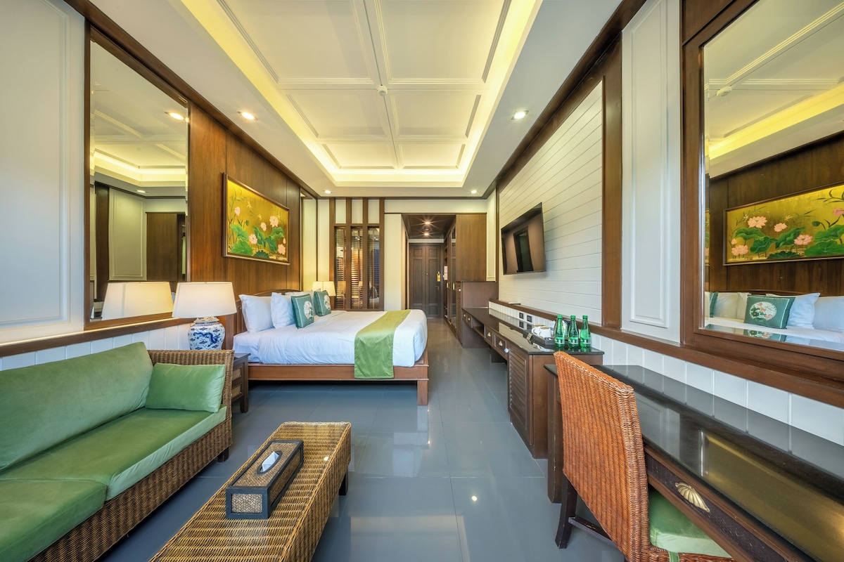 Habitación de hotel con cama, sofá verde, muebles de mimbre, un espejo y obras de arte.