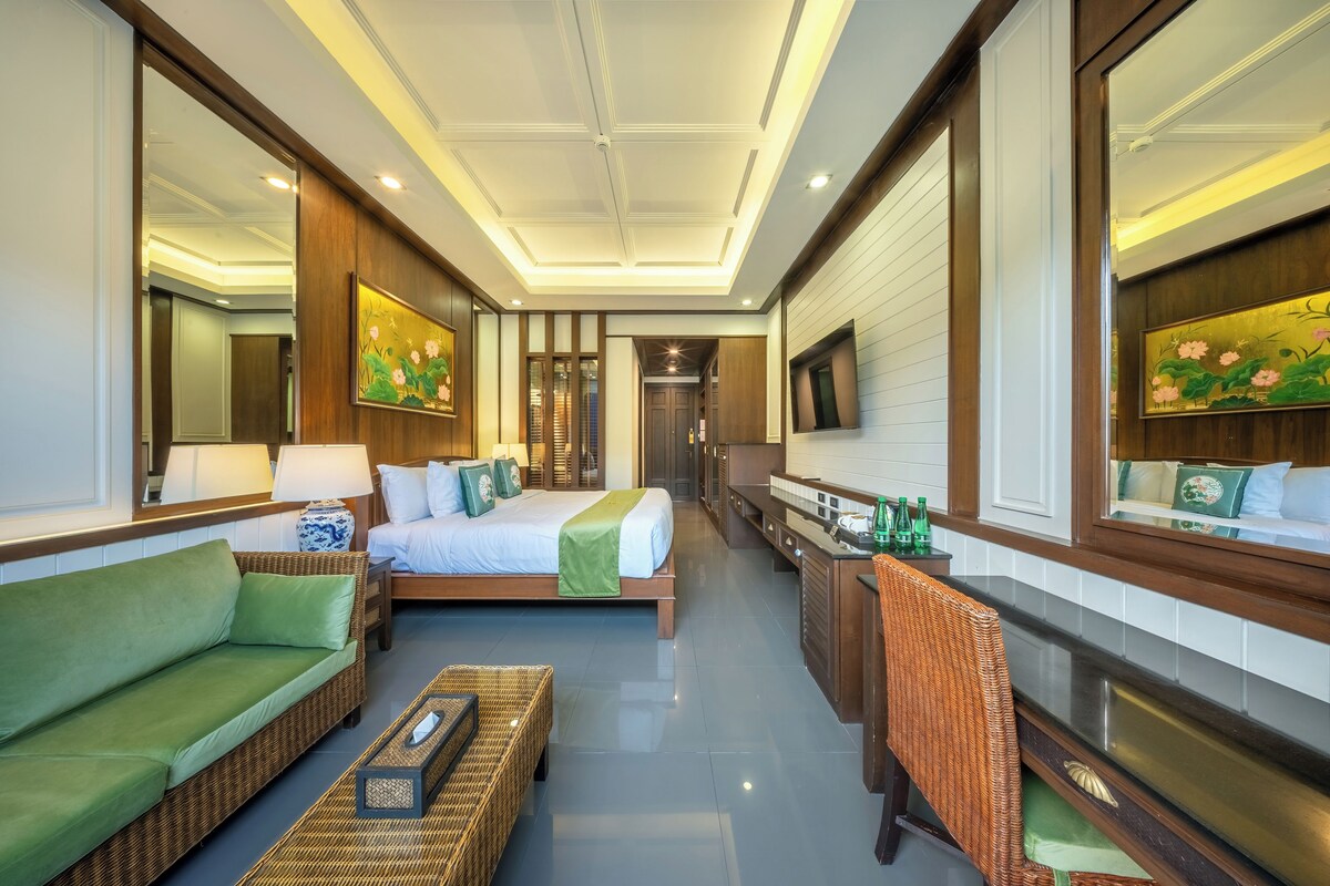 A hotel room with a bed, green sofa, wicker furniture, a mirror, and artwork.