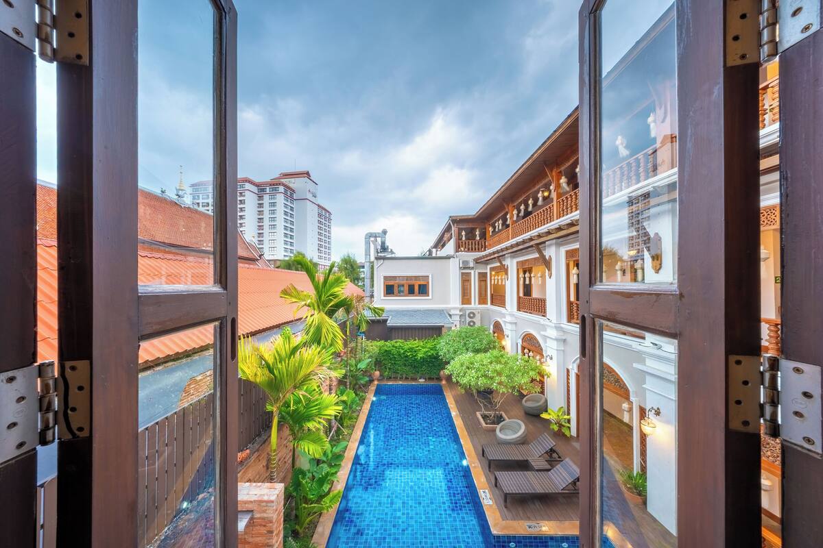 Une vue à travers une fenêtre donnant sur une piscine, entourée de verdure et bordée d’édifices à l’architecture traditionnelle.