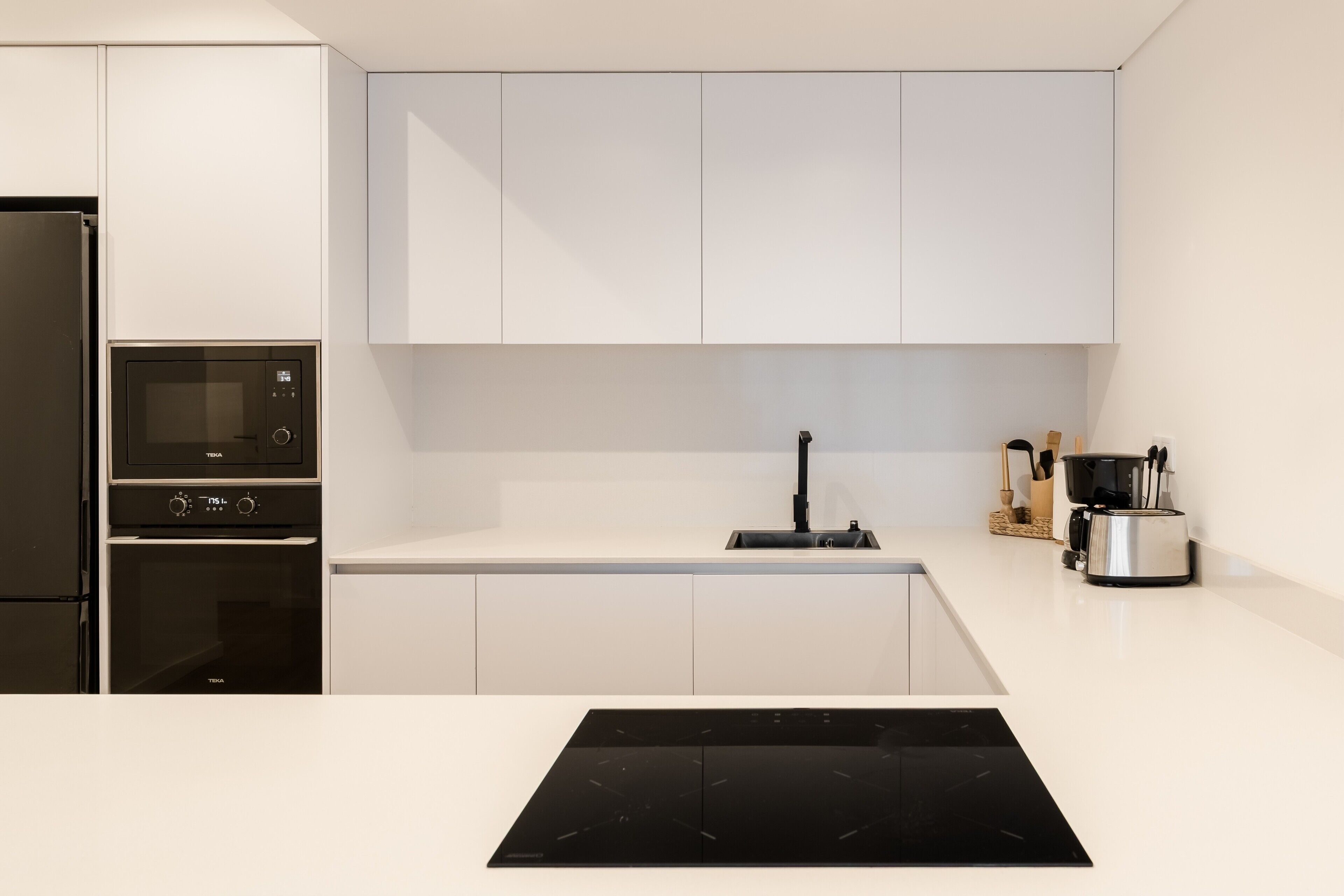 A modern kitchen with white cabinetry, a built-in oven, a microwave, a black induction cooktop, and a coffee maker on the countertop.
