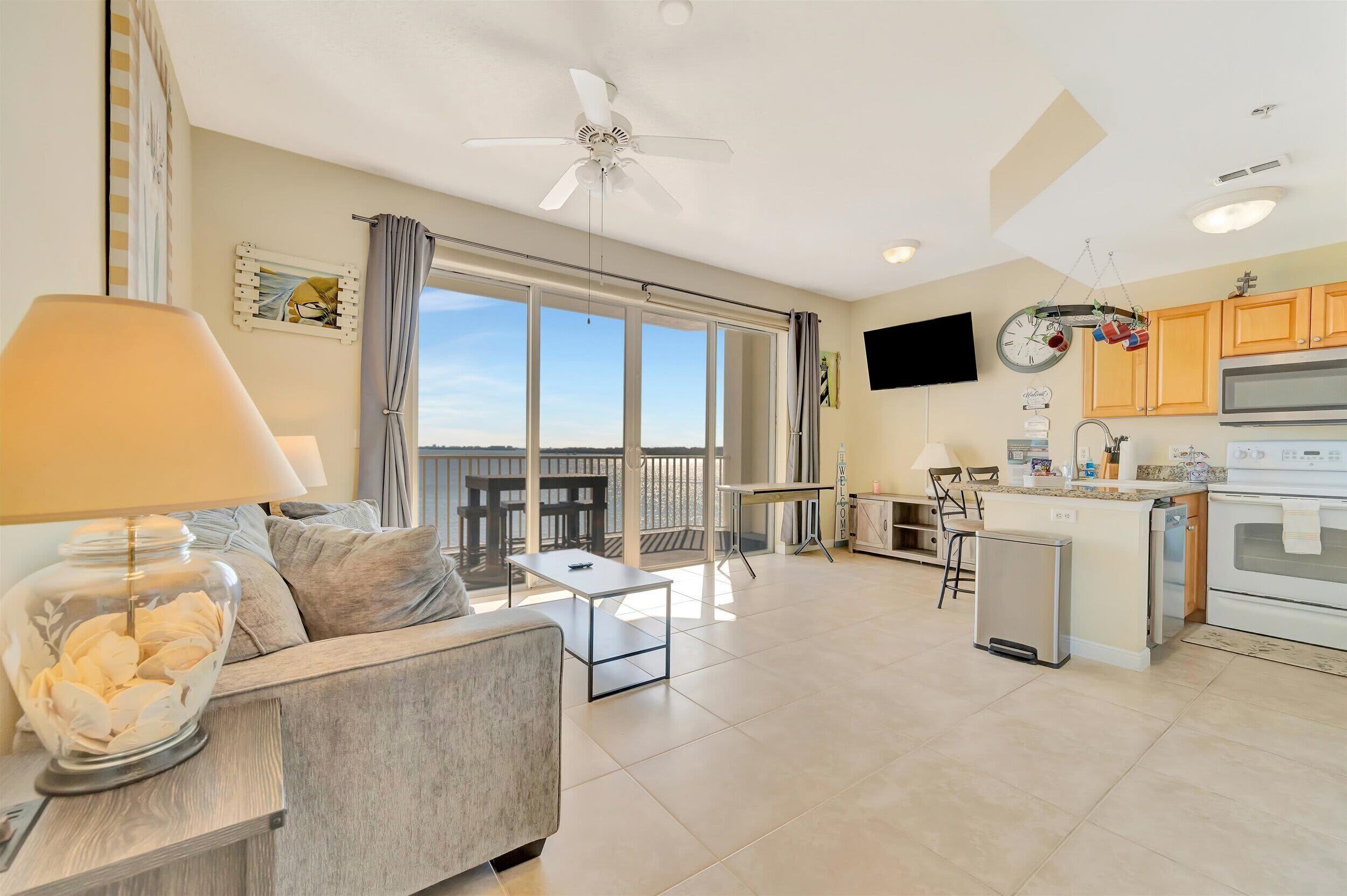 A spacious living area with a sofa, a coffee table, and a lamp. There is a balcony with a view, a kitchen with a refrigerator, and a television mounted on the wall.