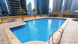 Ein Schwimmbecken mit einer Metallleiter, umgeben von einer gefliesten Terrasse und Palmen, mit Hochhäusern im Hintergrund.