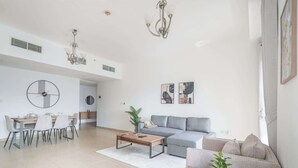 Ein großzügiger Wohnbereich mit grauem Sofa, einem Couchtisch und einem Esstisch für vier Personen. Zwei Wandleuchten und eine große runde Uhr sind vorhanden.