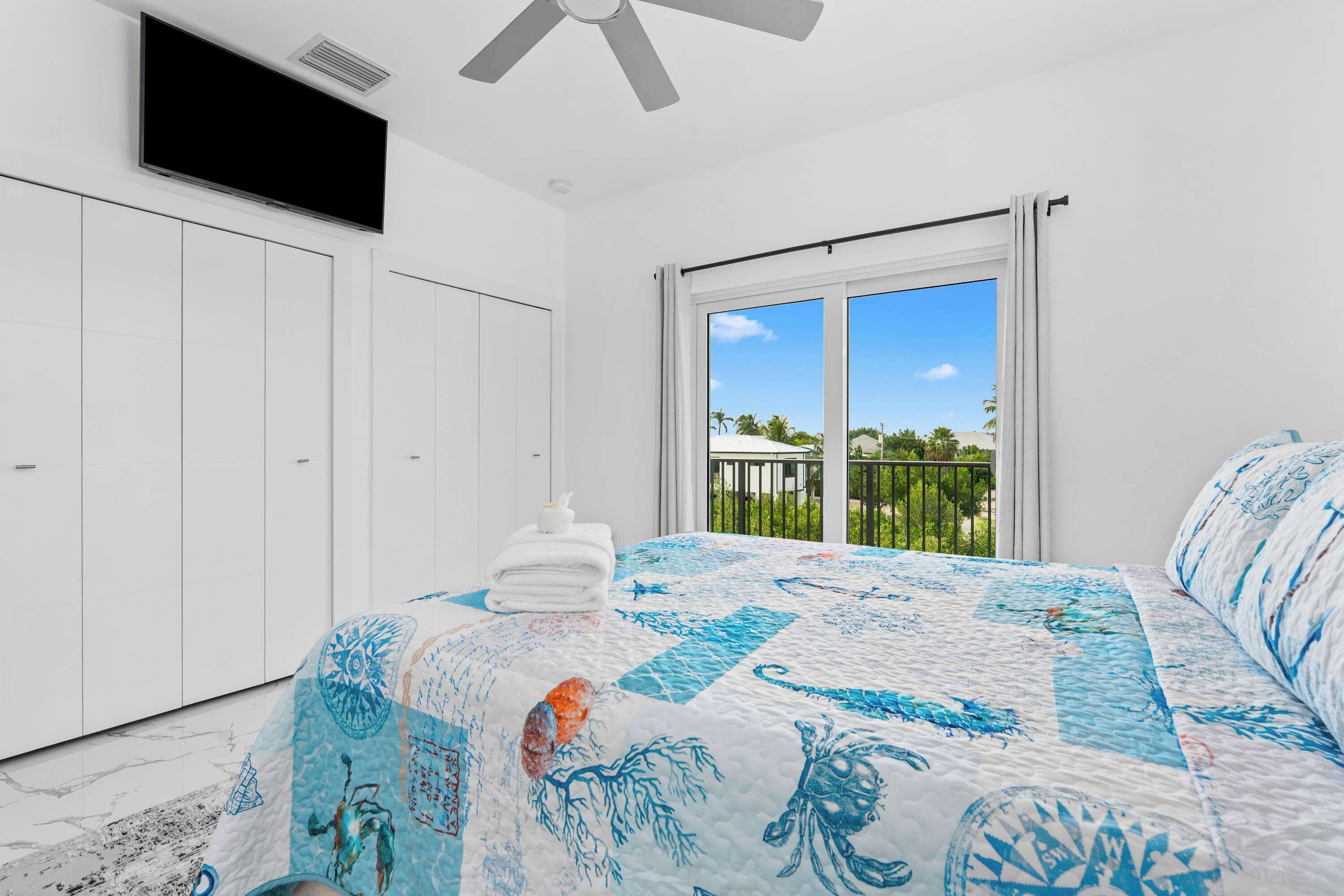 A bedroom with a large bed, a ceiling fan, a TV mounted on the wall, and a balcony with a view of palm trees.