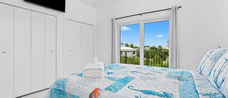 A bedroom with a large bed, a ceiling fan, a TV mounted on the wall, and a balcony with a view of palm trees.