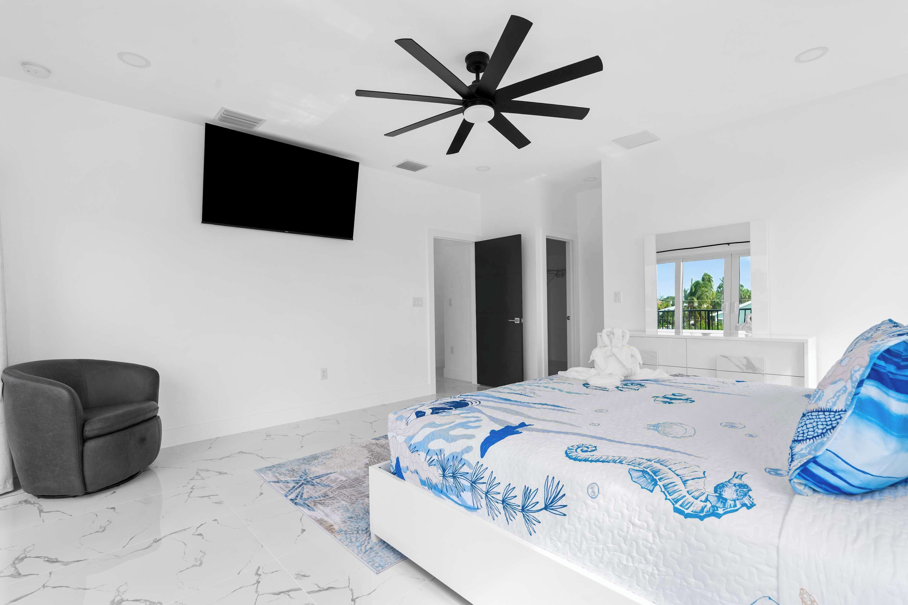 A modern bedroom with a bed, a ceiling fan, a flat-screen TV, and a view of the outdoors.
