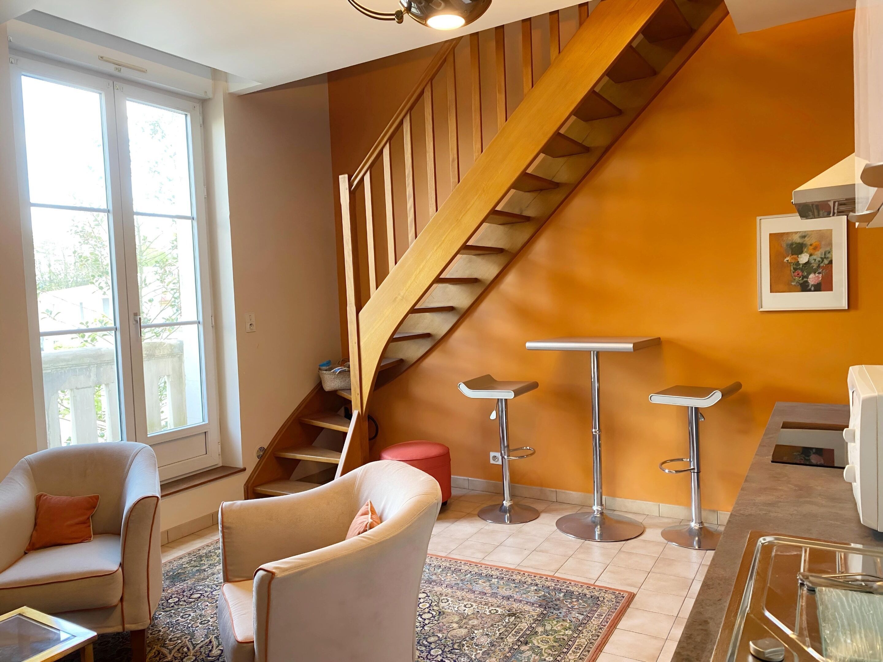 A modern living room with a staircase, two bar stools, a sofa, and a large window.