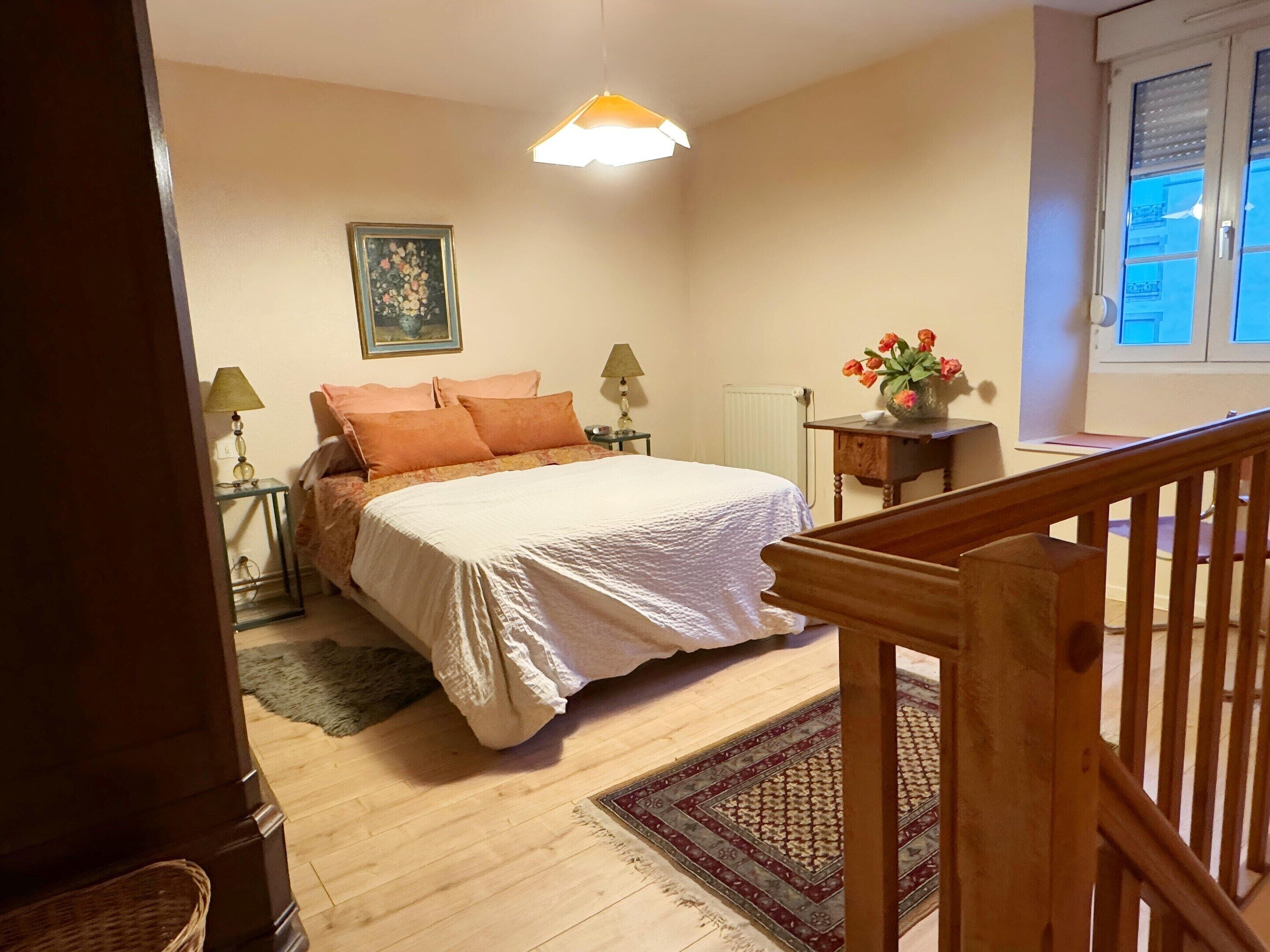 A bedroom with a bed, two bedside tables, a nightstand, a lamp, a painting, a window, and a wooden railing.