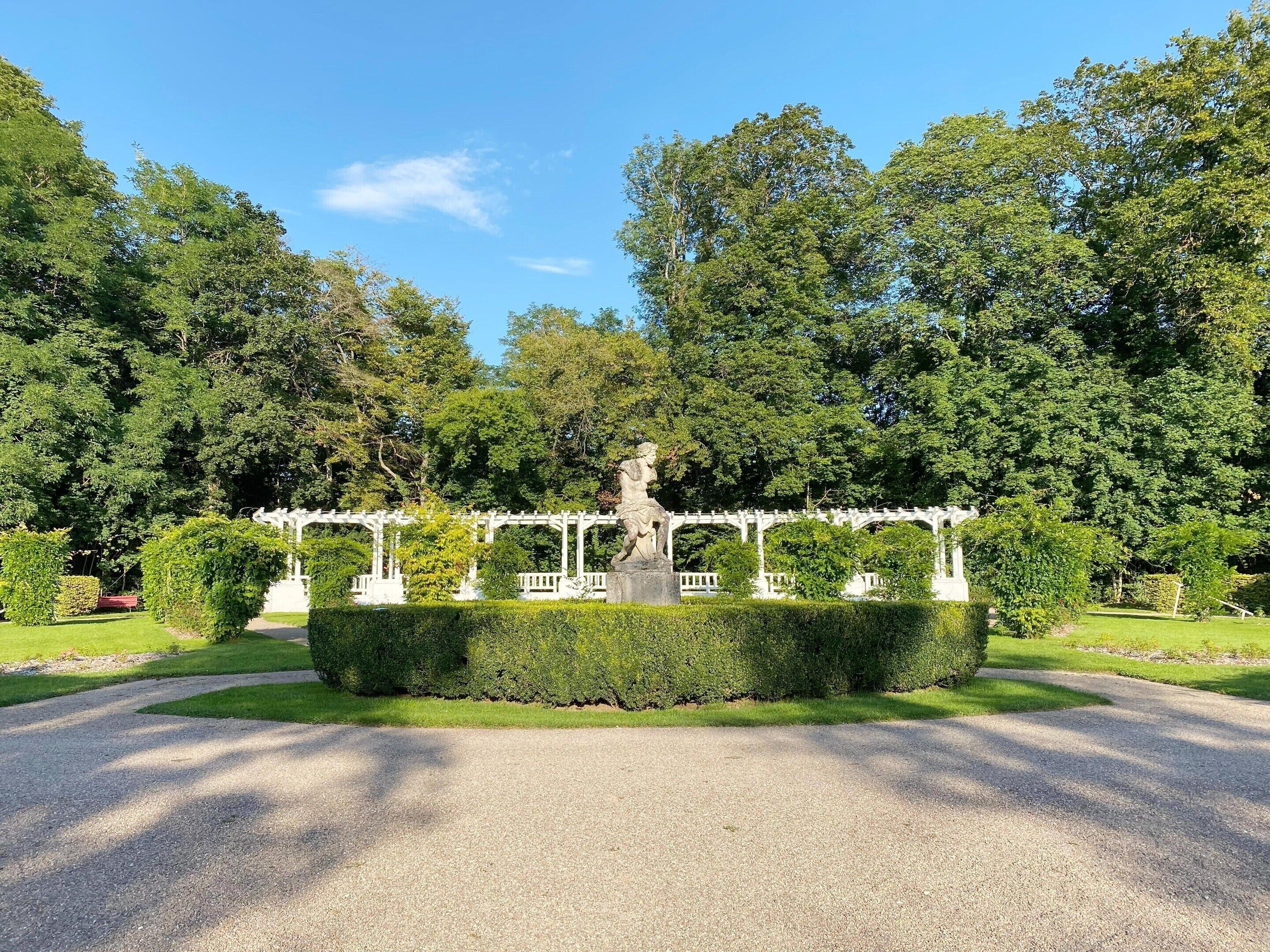 Een park met een standbeeld, een witte pergola en verzorgde hagen.