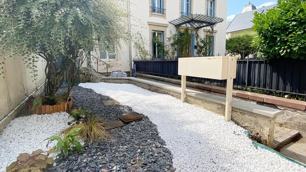 A courtyard with a gravel path, a wooden planter with plants, a metal pergola, and a building with a balcony in the background. - Amazing Holiday Home in the Heart of Vittel (Vittel)