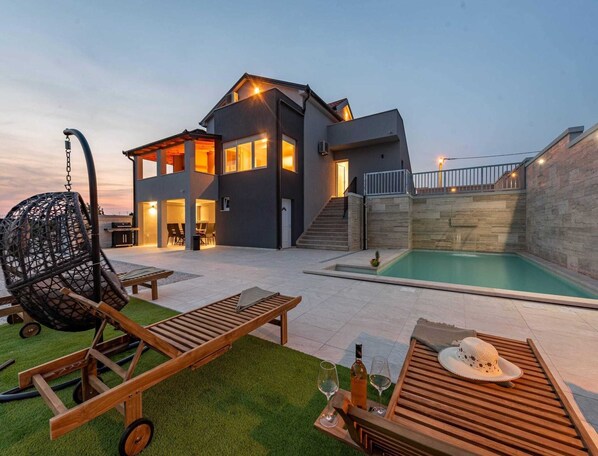 A modern house with a swimming pool, outdoor seating, and a hanging chair. - MY Dalmatia - Villa Pezer With Heated Pool & Sauna (Benkovac)