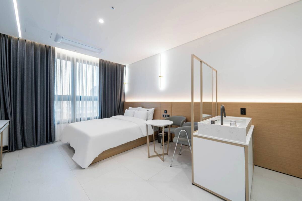 A modern hotel room with a bed, a small table, a chair, a desk, a mirror, and a bathroom with a sink.