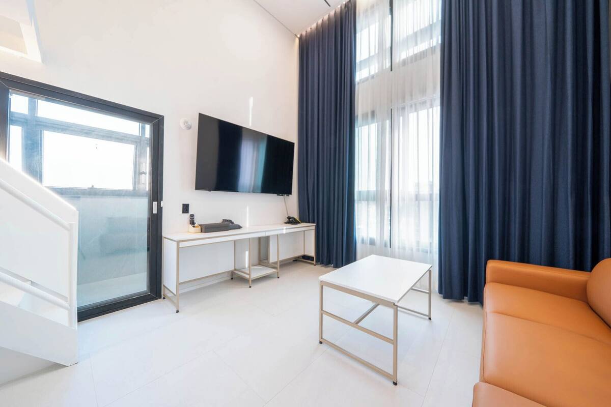 A modern living room with a flat-screen TV, a white desk, a small table, a sofa, and a sliding glass door leading to a balcony.