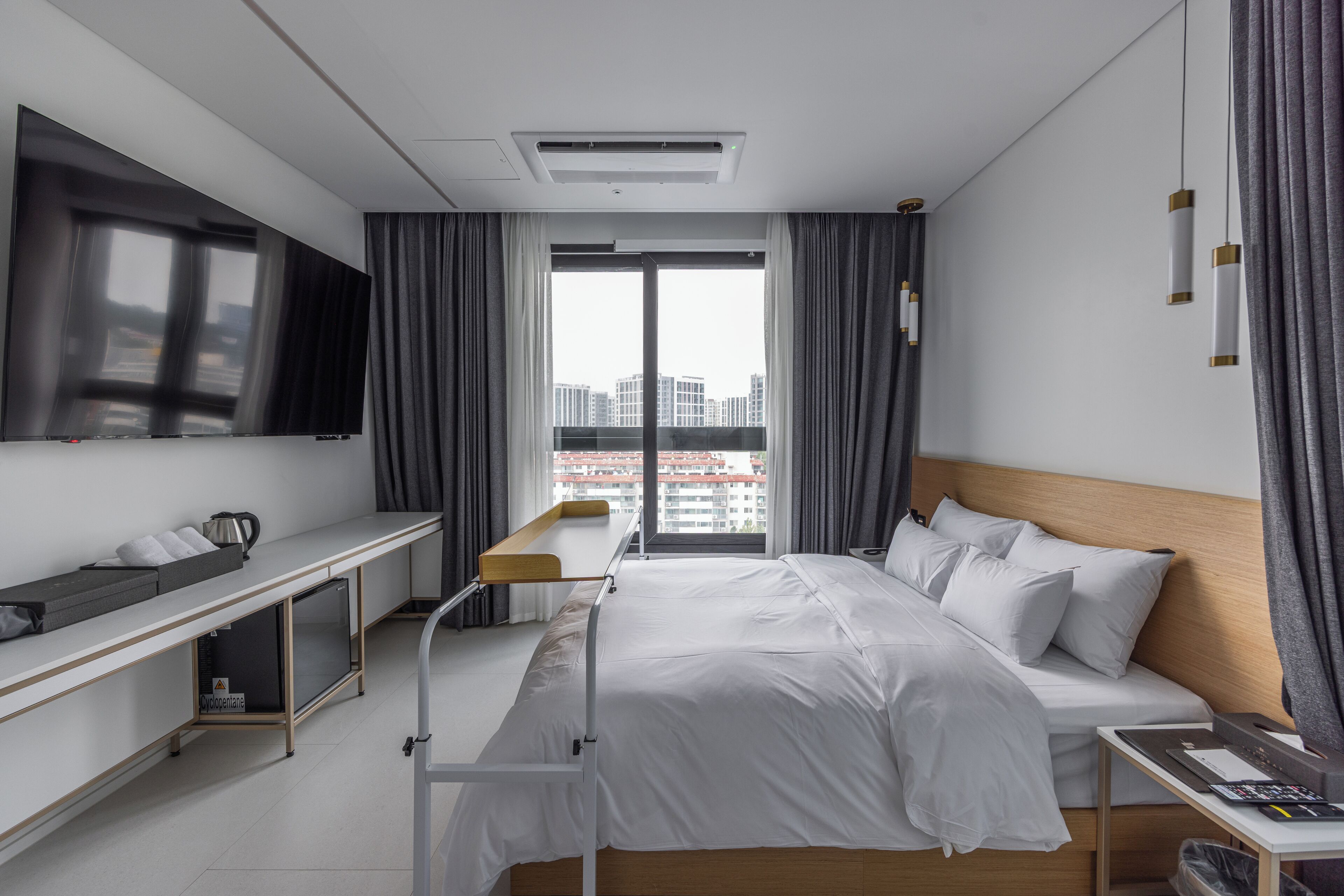 Una habitación de hotel moderna con cama, mesita de noche y un amplio ventanal con cortinas.