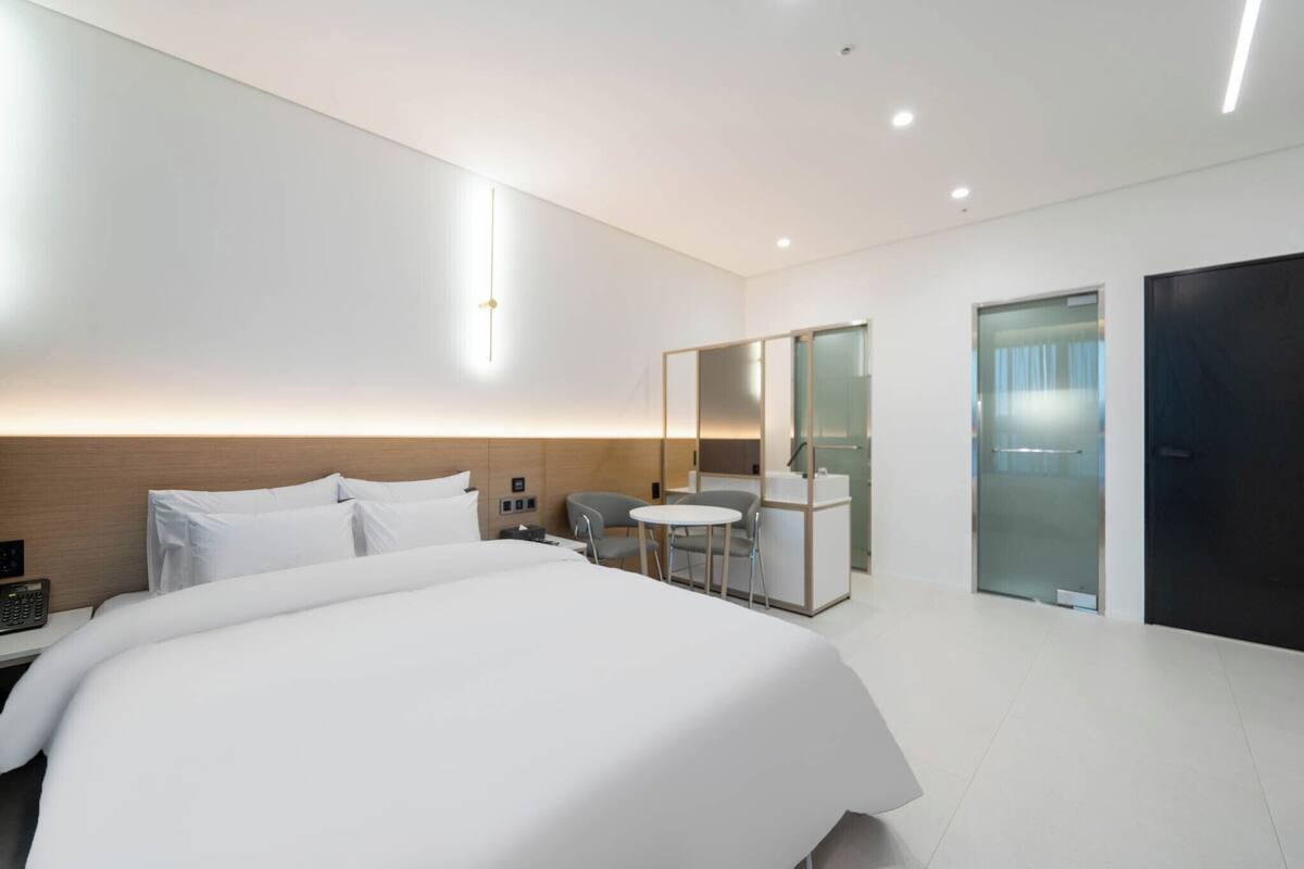 A modern hotel room with a large bed, a desk with a chair, a small round table, a mirror, and a bathroom with a glass door.