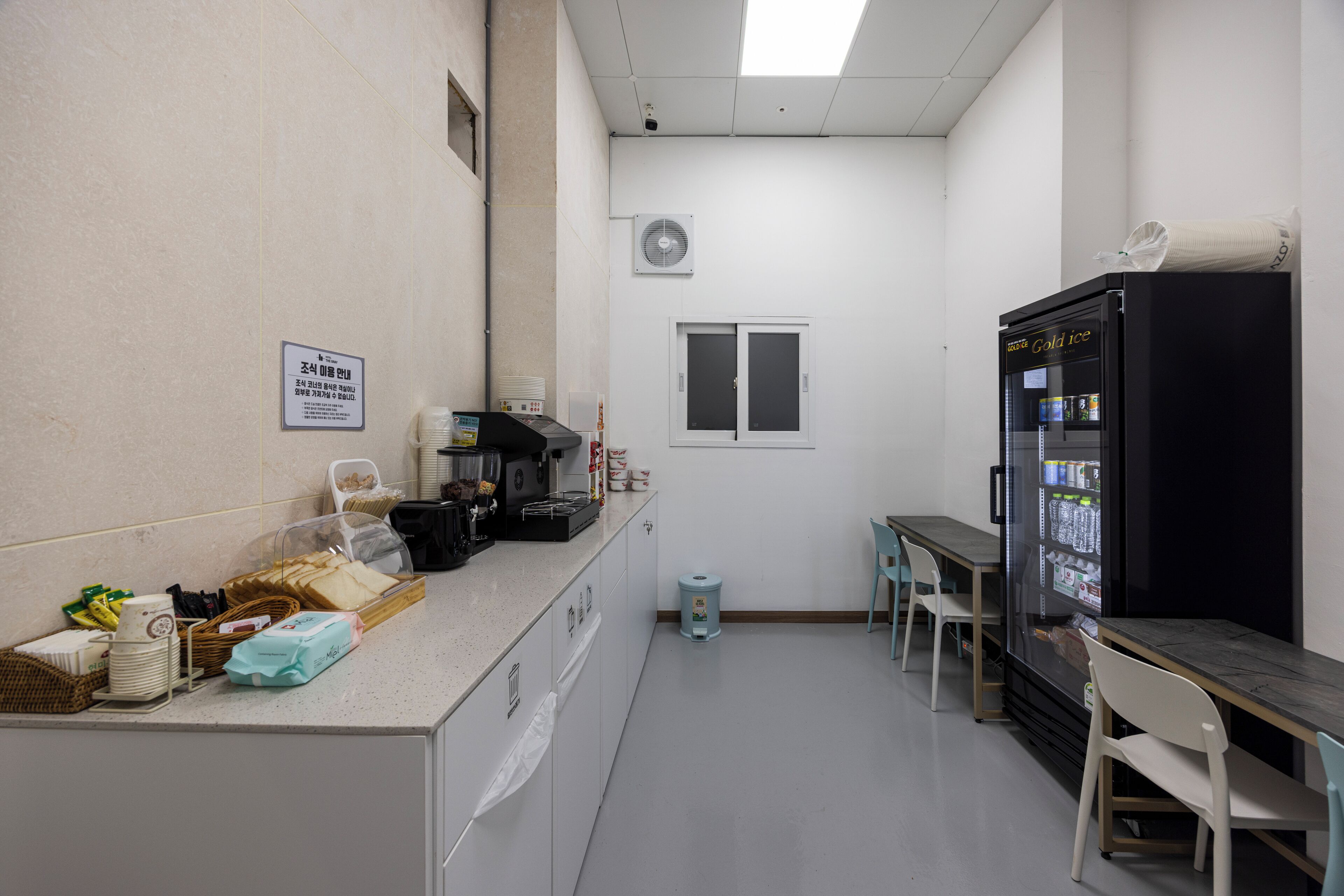 A kitchenette with a refrigerator, microwave, and coffee maker. There are chairs and a table, and a trash can is visible.