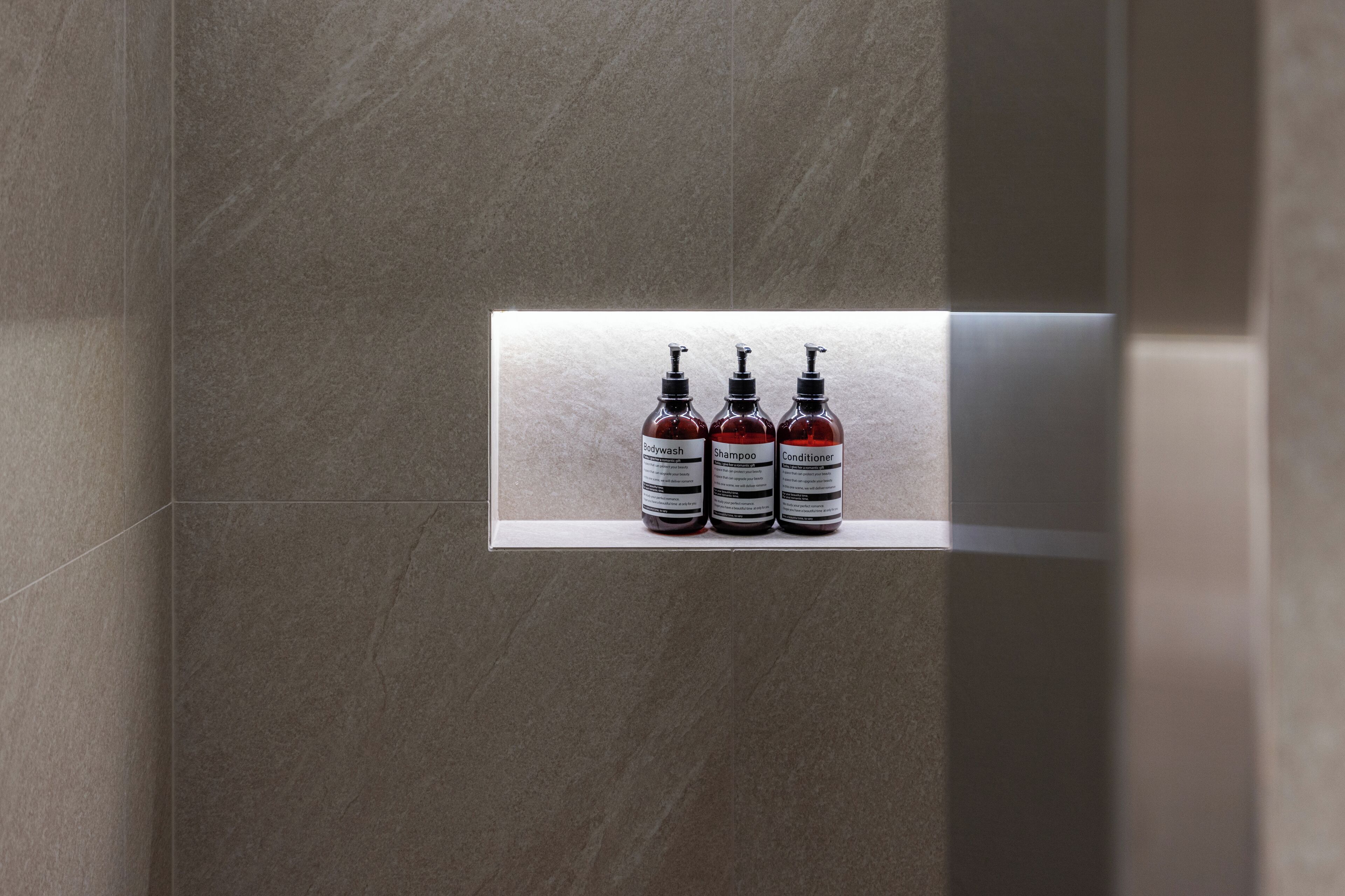 Een badkamer met drie flessen shampoo, conditioner en douchegel op een plank.